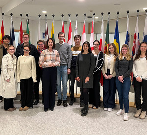 Gruppenfoto von Studierenden der HöV Rheinland-Pfalz mit Dr. Katarina Barley sowie den Dozierenden Dr. Anja Ehlers und Michael Eckhardt in einem Innenraum vor den Flaggen europäischer Staaten.