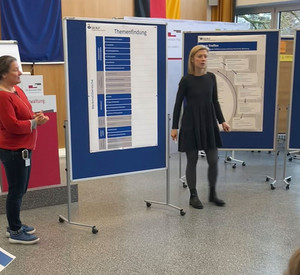 Kristin Flöck und Sonja Wittmann von der Unfallkasse Rheinland-Pfalz stehen in der Aula vor Studierenden. Im Hintergrund zu erkennen sind einige Stellwände.