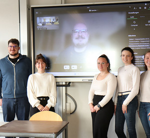 Gruppenfoto von Studierenden mit Dozent Frank Lenz in einem Seminarraum, im Hintergrund auf der Leinwand ist der digital zugeschaltete Referent Sören Damnitz von der Entwicklungsagentur Rheinland-Pfalz zu sehen.