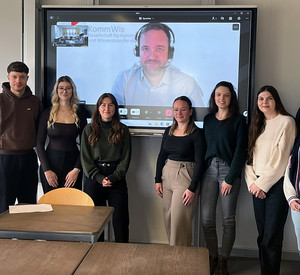 Studierende stehen in einem Seminarraum vor einem Bildschirm mit dem digital zugeschalteten Referenten Jan Druwe an der HöV RLP.