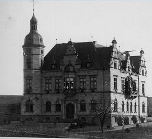Schwarzweißaufnahme eines historischen, repräsentativen Verwaltungsgebäudes mit Turm links, hohem Walmdach und reich verzierter Fassade, davor eine Straße und wenige kahle Bäume in offener Landschaft.