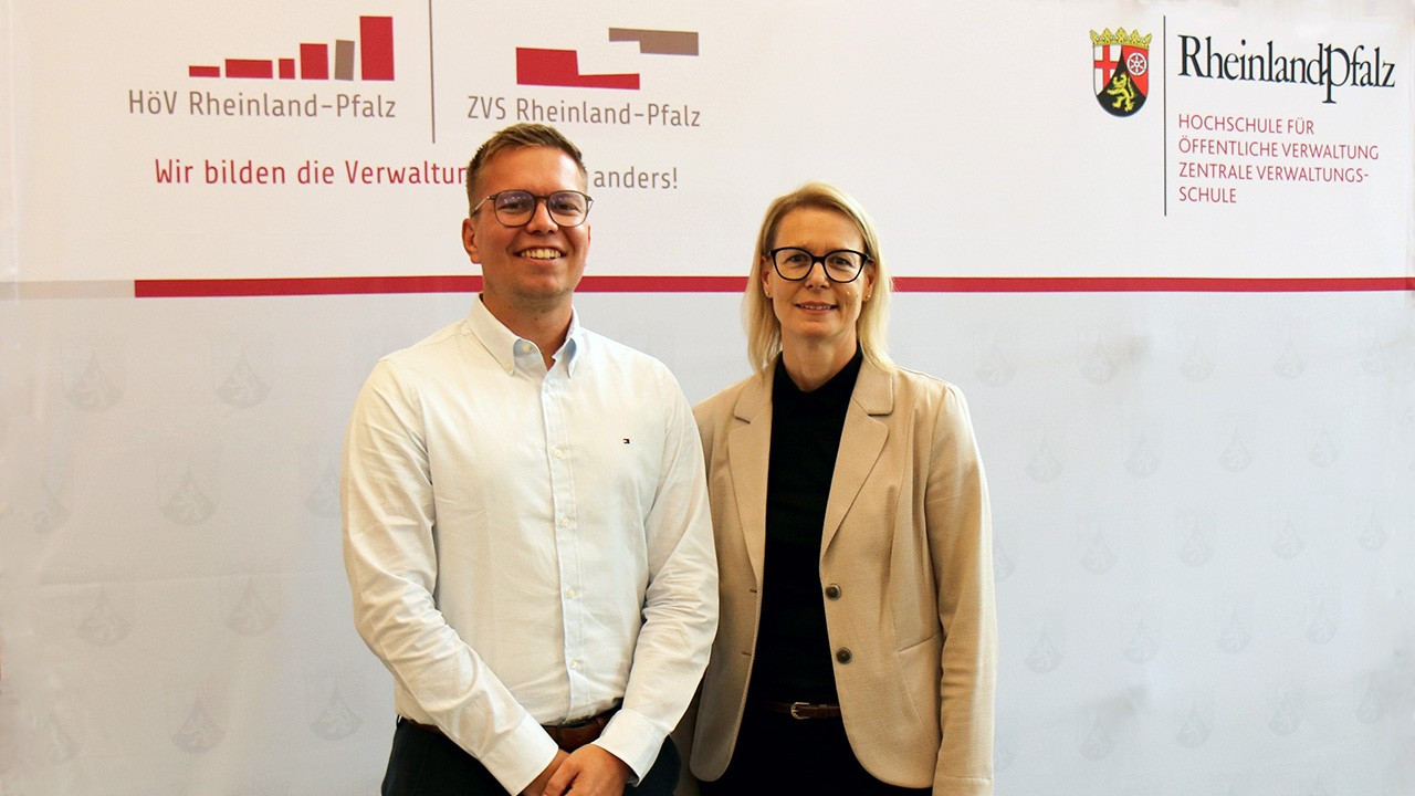 Direktorin Alice Schmidt und Referent Julian Schmitz stehen vor der Pressewand der Hochschule für öffentliche Verwaltung Rheinland-Pfalz bei der Infoveranstaltung zur Beihilfe.