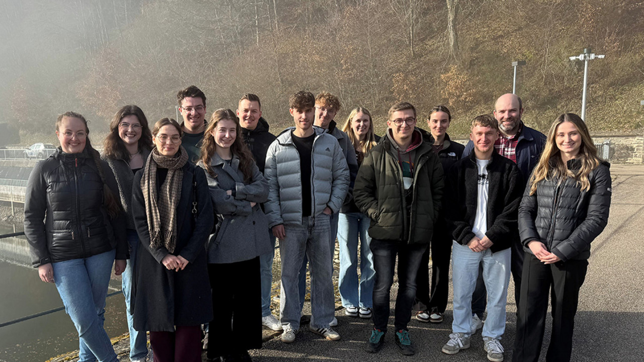 Eine Gruppe von Studierenden steht gemeinsam mit Dozent Markus Stein im Freien an der Riveristalsperre bei Trier. Im Hintergrund sind Wasser, Uferbereich und bewaldeter Hang zu sehen. 
