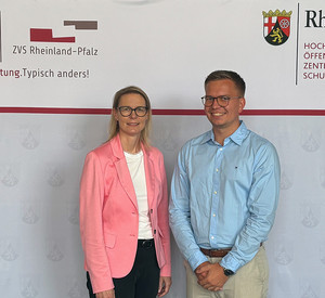 Direktorin Alice Schmidt im rosafarbenen Blazer und Julian Schmitz im hellblauen Hemd stehen vor der Pressewand der HöV Rheinland-Pfalz und ZVS Rheinland-Pfalz. Beide schauen lächelnd in die Kamera.