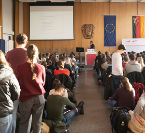 Ein Hörsaal an der HöV RLP in Mayen. Vorne steht ein Referent und spricht zu vielen Studierenden. Auf der Leinwand ist eine Präsentationsfolie mit dem Titel „Verfassungsschutz Rheinland-Pfalz“ zu sehen.