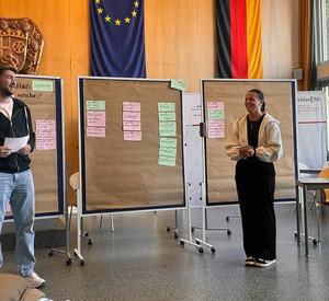 : Zwei Studierende stehen vor Pinnwänden mit rosa und grünen Moderationskarten und moderieren ein Gespräch. Auf einer Pinnwand steht die Frage: „Welche Aspekte sollten bei einer Reise beachtet werden?“