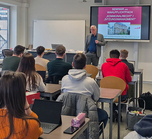 Bürgermeister Uwe Bruchhäuser spricht in einem Seminarraum in Mayen mit Studierenden der HöV RLP und Lehrgangsteilnehmenden der ZVS RLP. Im Hintergrund ist ein Whiteboard mit einer Präsentation zu erkennen.