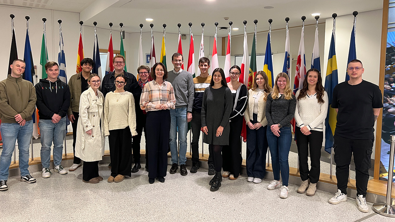 Gruppenfoto von Studierenden der HöV Rheinland-Pfalz mit Dr. Katarina Barley sowie den Dozierenden Dr. Anja Ehlers und Michael Eckhardt in einem Innenraum vor den Flaggen europäischer Staaten.