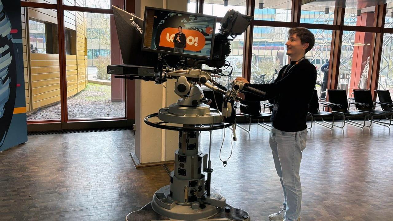 Ein Studierender der HöV Rheinland-Pfalz steht im ZDF an einer Studiokamera und blickt auf einen Monitor mit seinem Bild. 