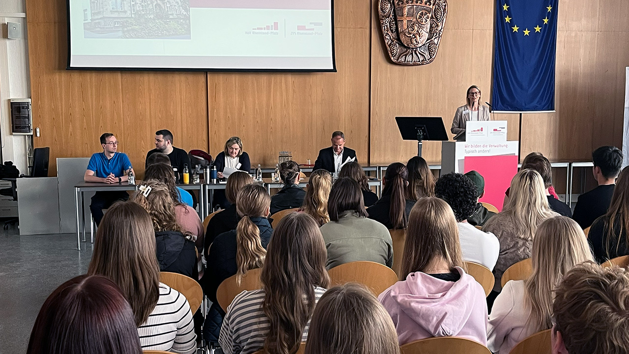 Die Direktorin begrüßt Teilnehmende des Hochschulinformationstags in der Aula der HöV RLP und ZVS RLP in Mayen. Vorne sitzen weitere Personen an einem Tisch.