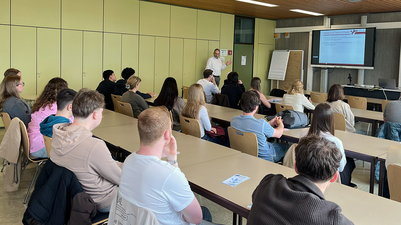Teilnehmende sitzen in einem Lehrsaal und hören einem Vortrag zu. Auf einem Bildschirm sind Informationen zu Theorie und Praxis zu sehen.