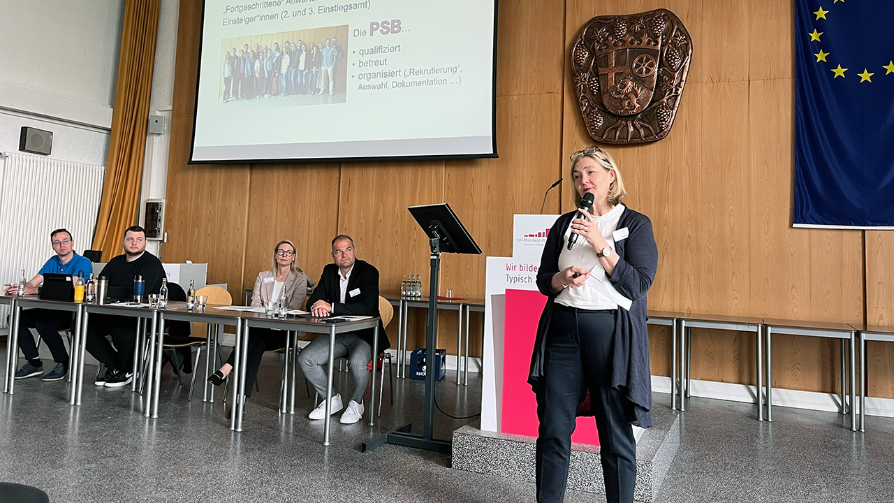 Eine Person spricht mit Mikrofon in der Aula vor Teilnehmenden. Auf der Leinwand sind Informationen zur Psychosozialen Beratungsstelle zu sehen.